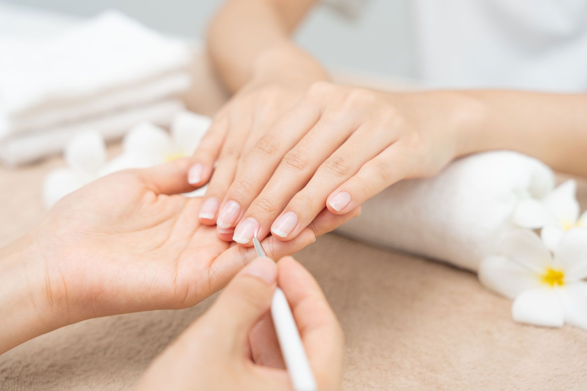 A manicurist uses a cuticle pusher on a client's fingernail, with white flowers nearby on a neutral surface.