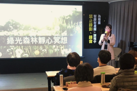 一位女士正在演講，她身後的螢幕上顯示著「綠色森林冥想」。聽眾們圍坐在桌旁。