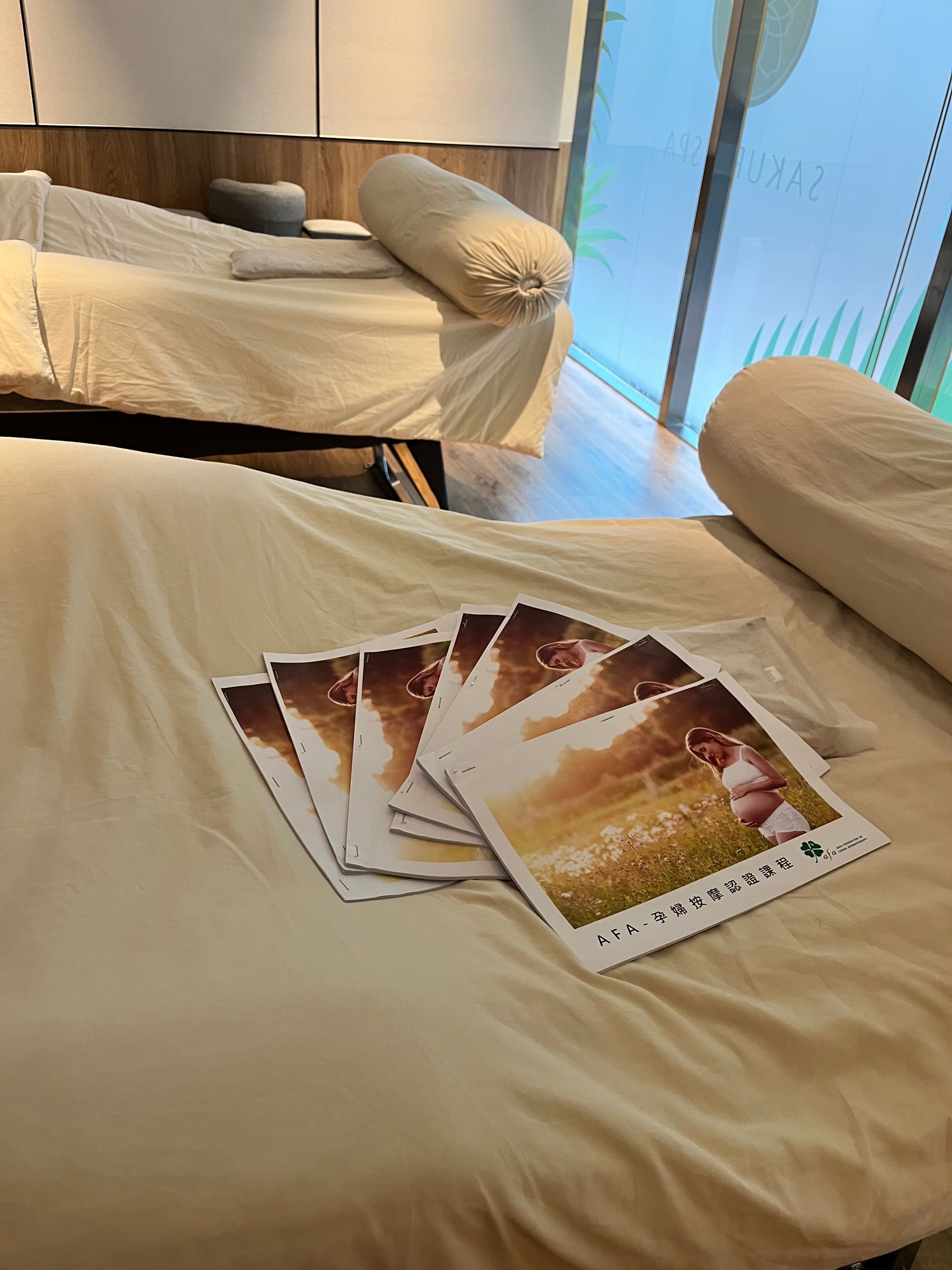米色床上堆放著一疊宣傳冊,旁邊放著一個長枕頭,背景還有一張床。窗外透進光線。