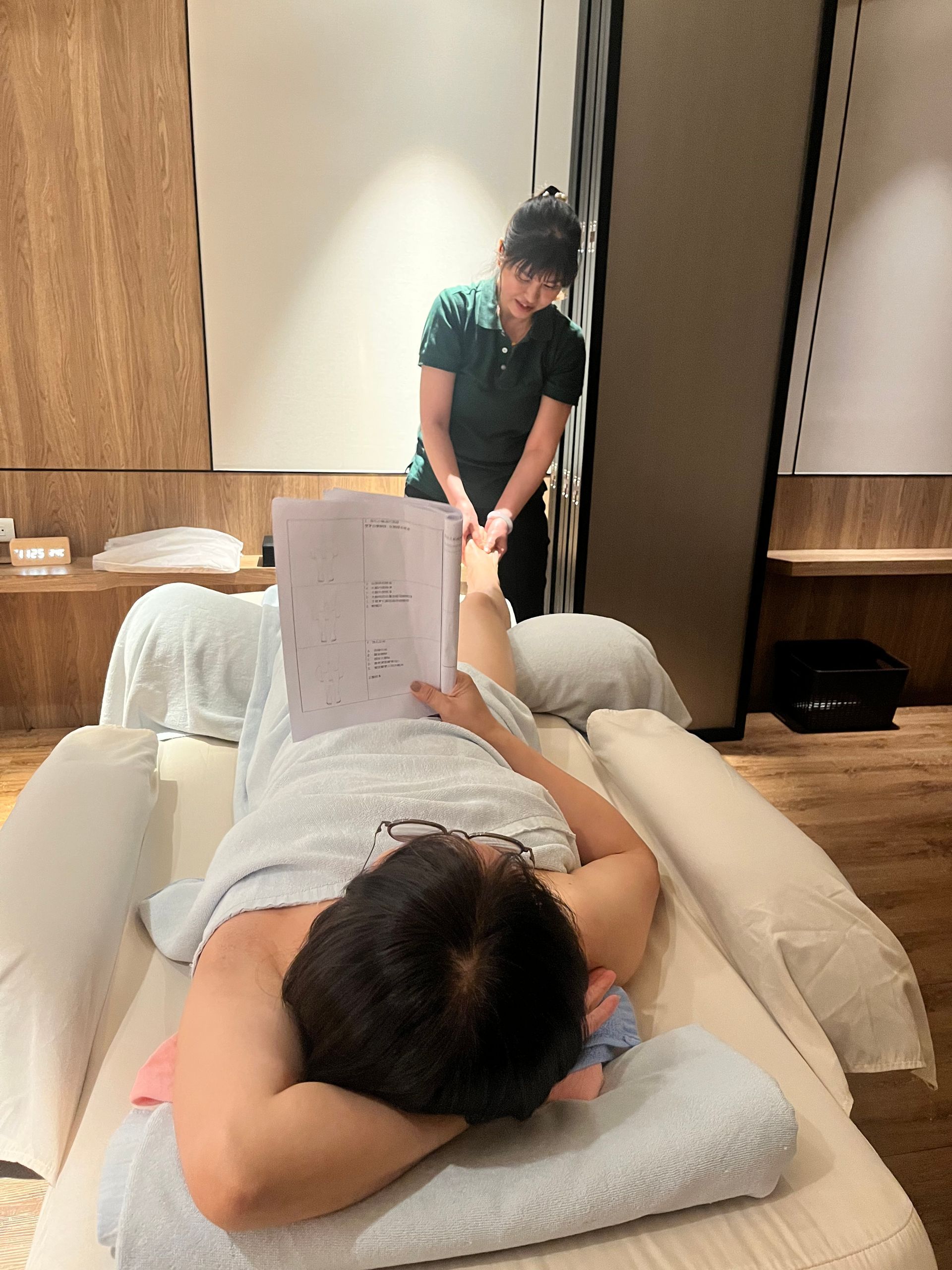 在水療中心接受足部按摩的人；身穿綠色襯衫的按摩師，正在閱讀，燈光柔和。