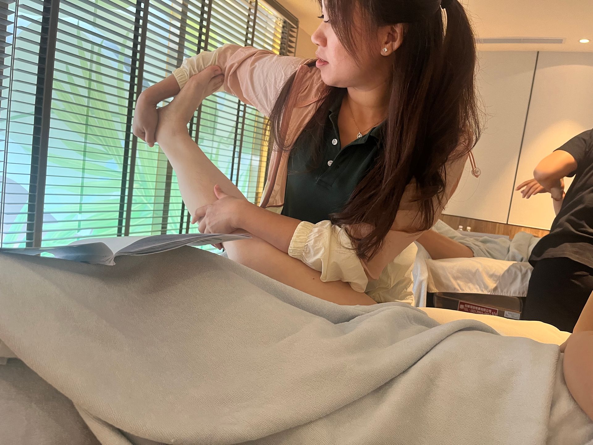 一位女士正在按摩,彎曲著手臂。按摩者躺在按摩床上。房間光線明亮,呈綠褐色調,窗簾拉上了。