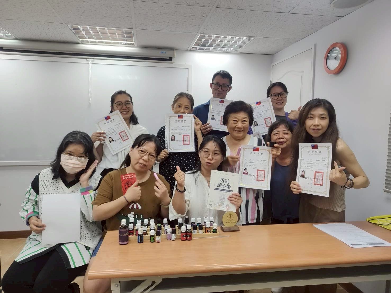一群人在教室裡，手持證書，微笑地圍著擺放著精油瓶的桌子合照。