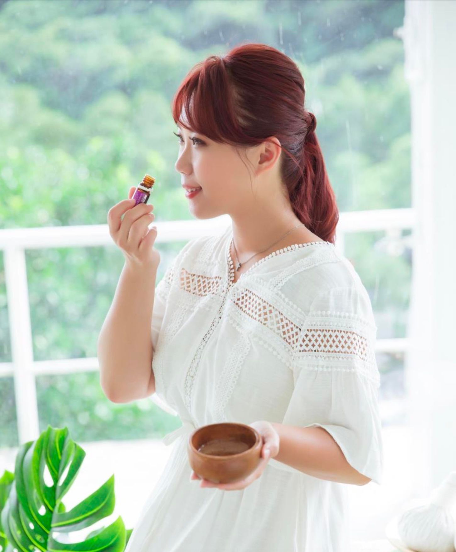 身穿白色洋裝的女子，手持木碗，在戶外綠樹成蔭的地方，聞著一個小瓶子的味道。