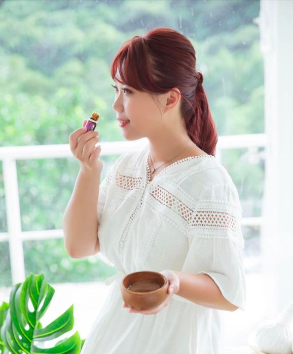 身穿白色洋裝的女子，手持木碗，在戶外綠樹成蔭的地方，聞著一個小瓶子的味道。