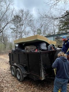 Two people loading a black trailer with bags and a mattress in a wooded area