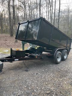 Black dump trailer tilted forward on a gravel road in a wooded area.