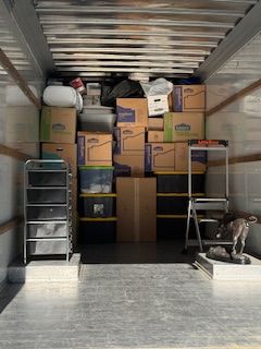 Packed moving truck with stacked boxes, shelves, and a ladder inside, viewed from the open rear.
