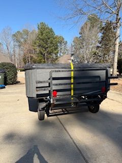 Gray dump trailer with a black frame and yellow tie-down strap parked on a driveway