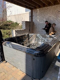 Outdoor hot tub on a patio with clear cover, beside a brick wall and two people nearby.