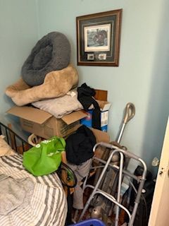 Cluttered corner with stacked boxes, a walker, and pet beds beside a framed wall picture