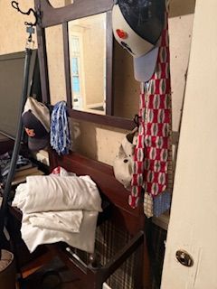 Bedroom vanity area with mirror, hanging clothes, folded towels, and a jacket draped over a chair.
