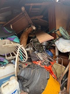 Cluttered storage space with piled furniture, chairs, boxes, tools, and miscellaneous household items.