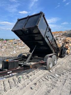 Black dump trailer tipped up at a landfill, unloading debris under a blue sky.