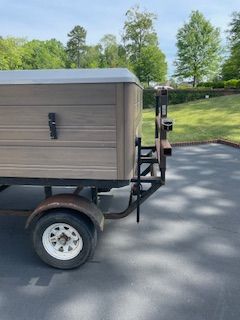 Small gray utility trailer with a single wheel, parked on a paved road beside grass and trees.