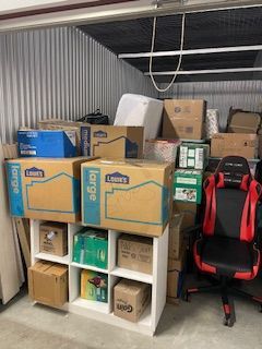 Storage shelf with stacked cardboard boxes and a red-and-black gaming chair in a cluttered garage attic area