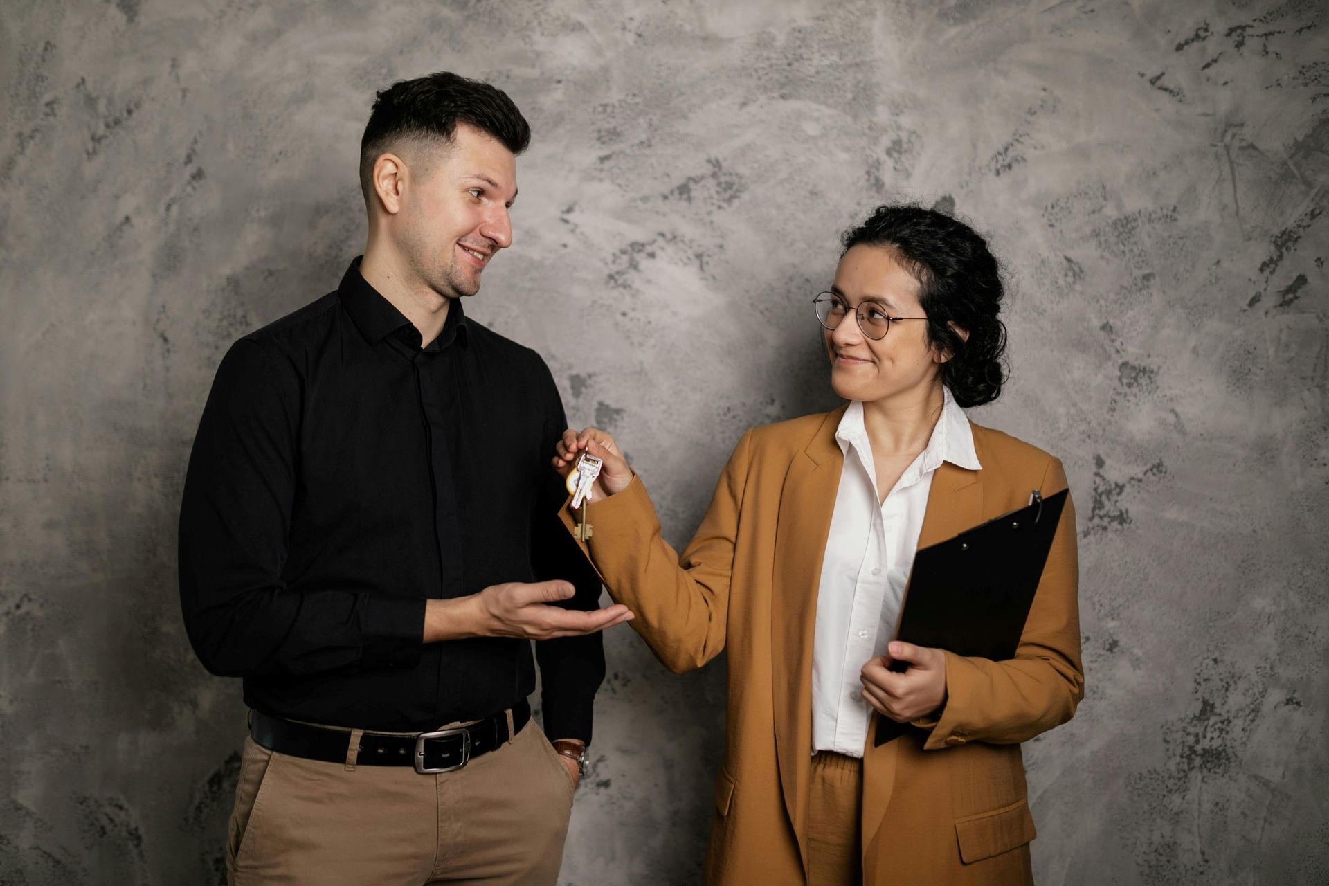 A woman in a tan blazer hands a key to a man in a black shirt against a grey textured wall.