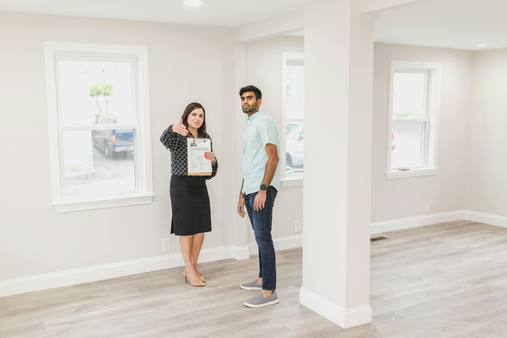 A person holding a clipboard points toward a window as another person looks on in a bright, empty room with wood flooring.