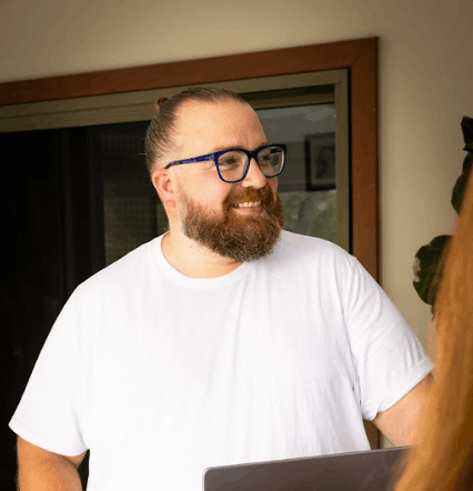 Man with glasses and beard smiles, looks right. Wearing white tee, hair in bun. Indoors near door.