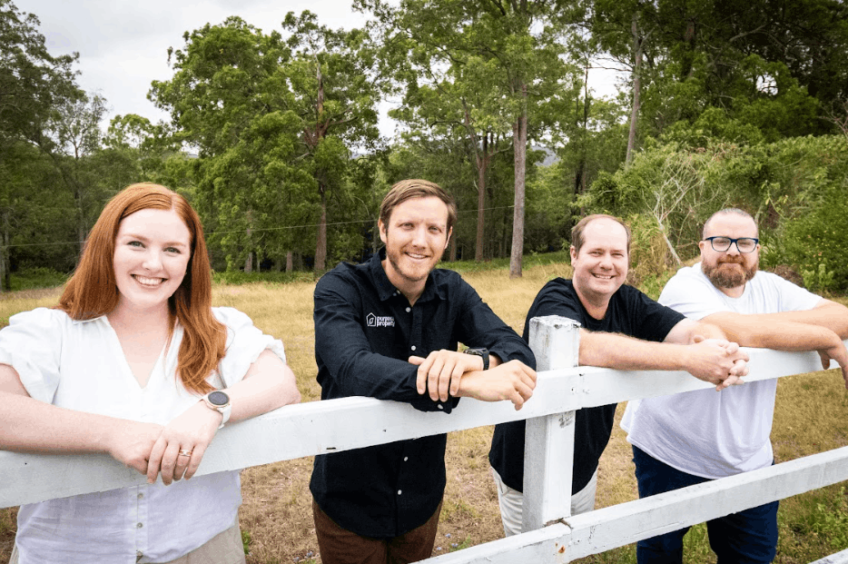 Four people lean on a white fence in a grassy field, smiling at the camera. Trees form a backdrop.