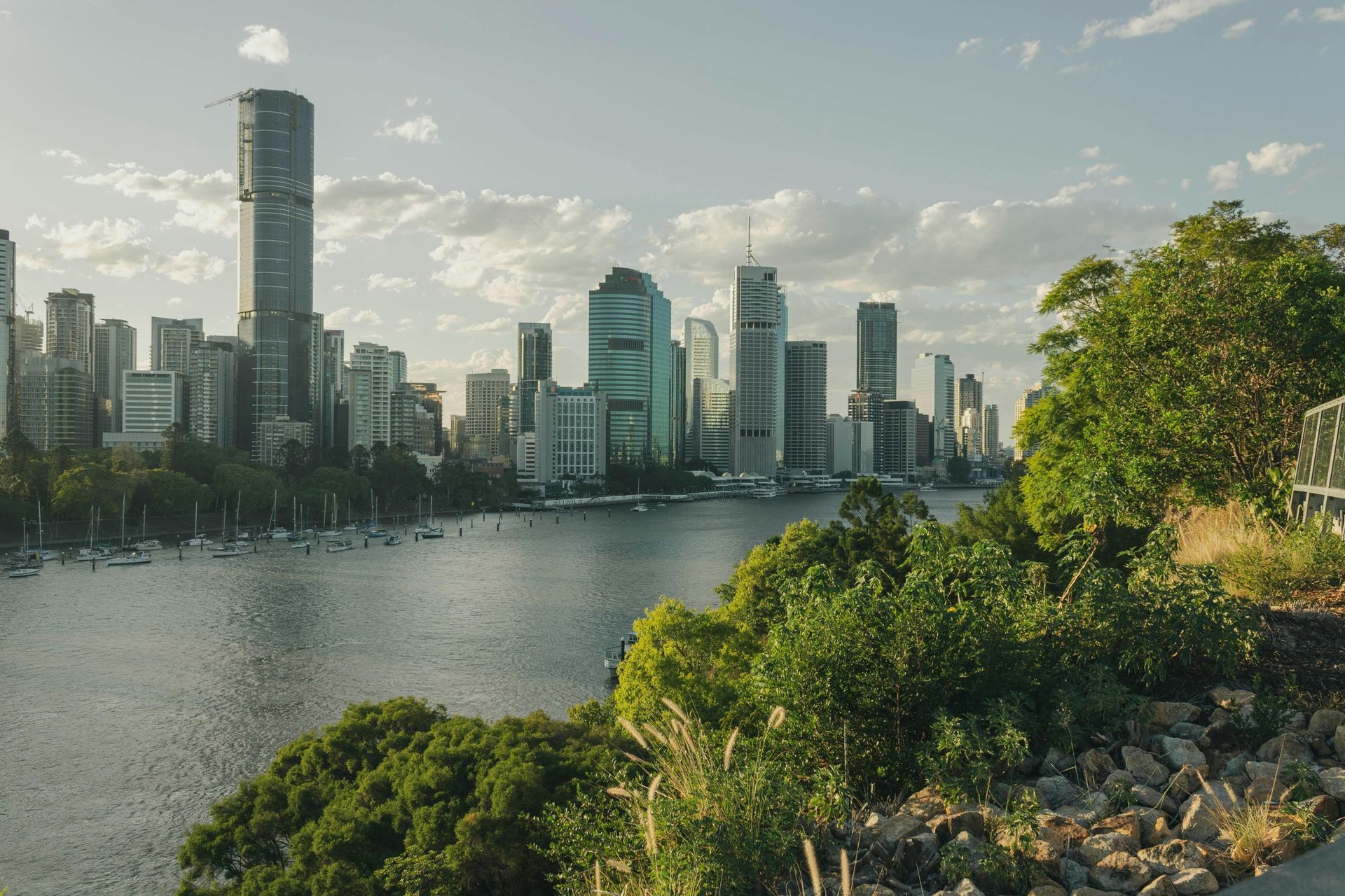 Brisbane city skyline and river at sunset, framed by lush green trees in the foreground.