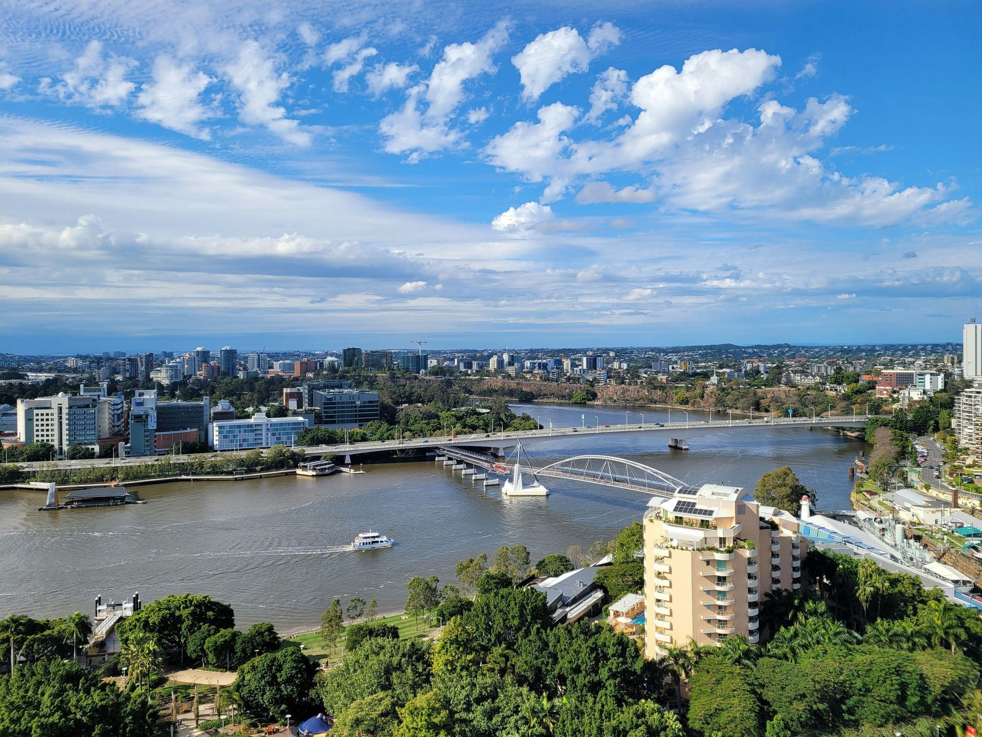 A high-angle view of a river winding through a cityscape under a bright blue, cloudy sky.