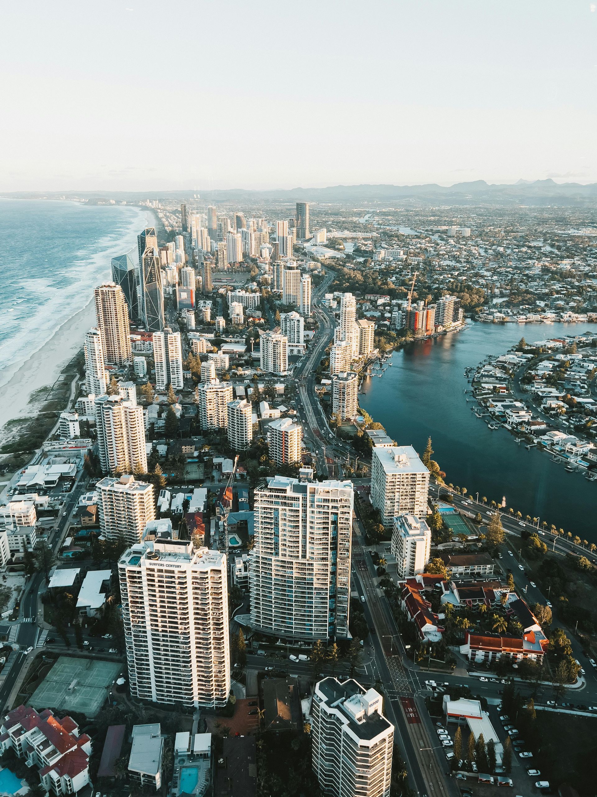 Aerial view of a coastal city with high-rise buildings along a beach and a winding river under a hazy, warm sky.