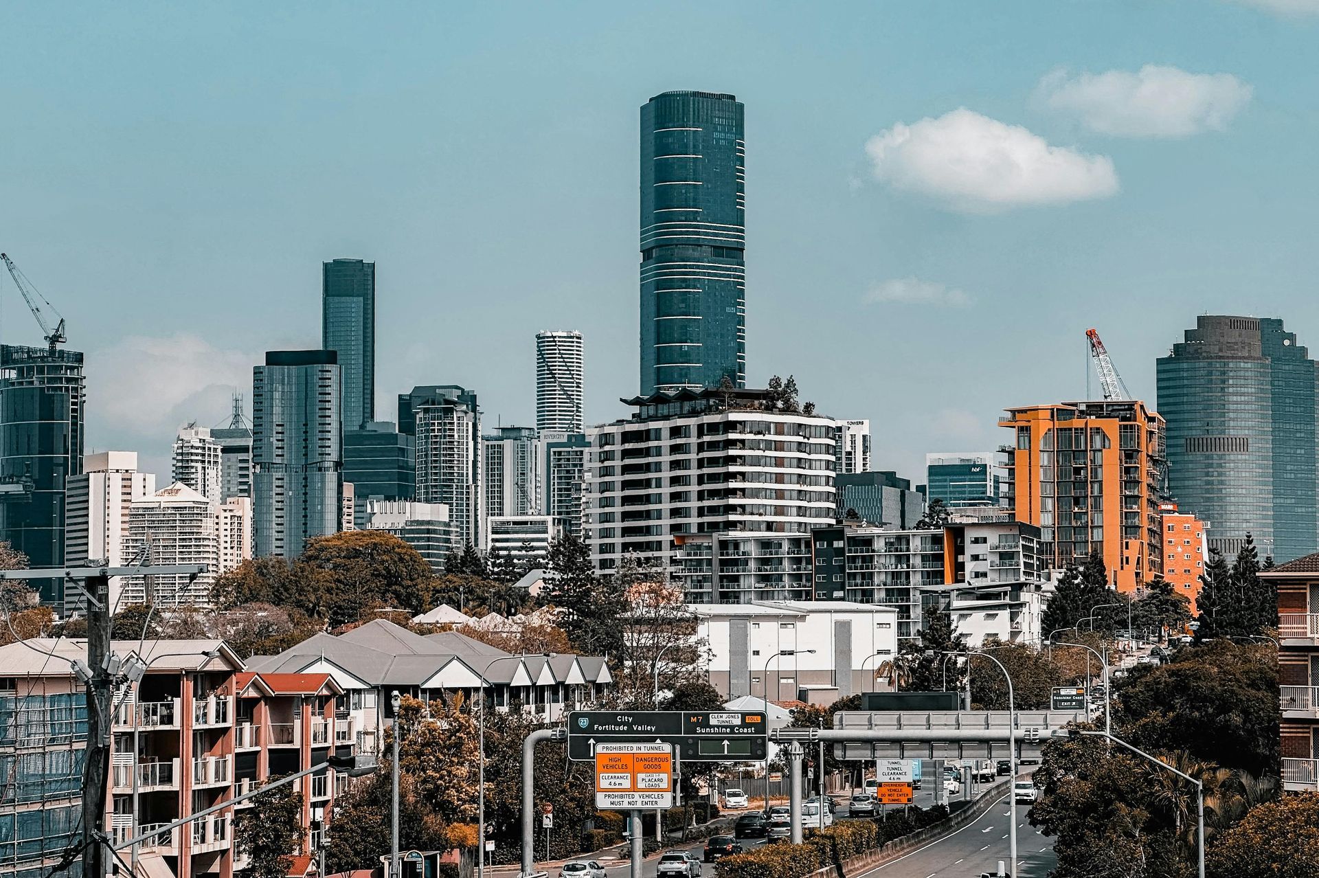A modern city skyline featuring a prominent dark skyscraper rising above smaller buildings and lush trees in the foreground.