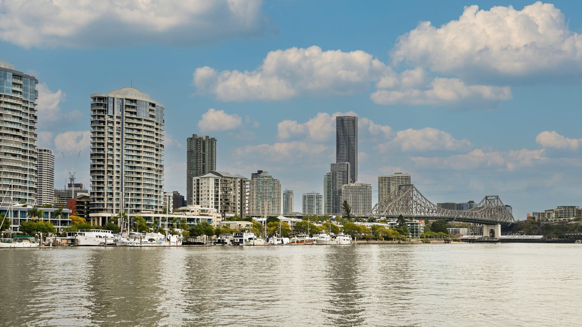 A riverfront cityscape with high-rise residential buildings, lush greenery along the bank, and a bridge under a blue sky.