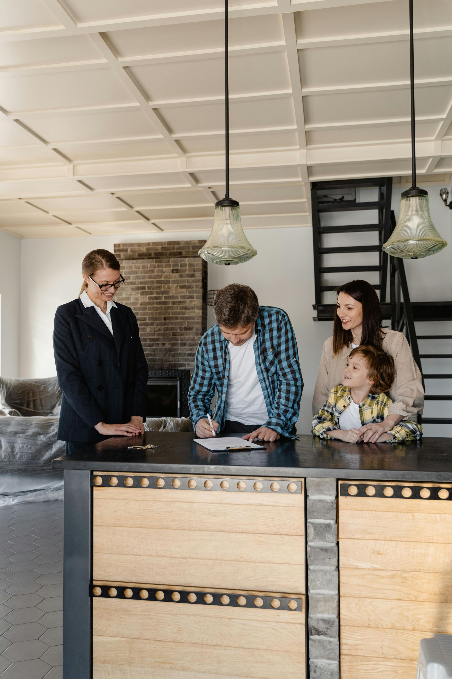 A person in a blazer observes a man signing documents at a kitchen island while a woman and child stand beside him.