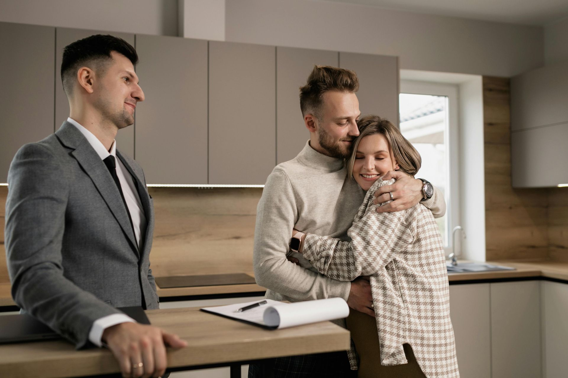 A real estate agent stands by a counter as a couple embraces in a modern kitchen, celebrating a home purchase.