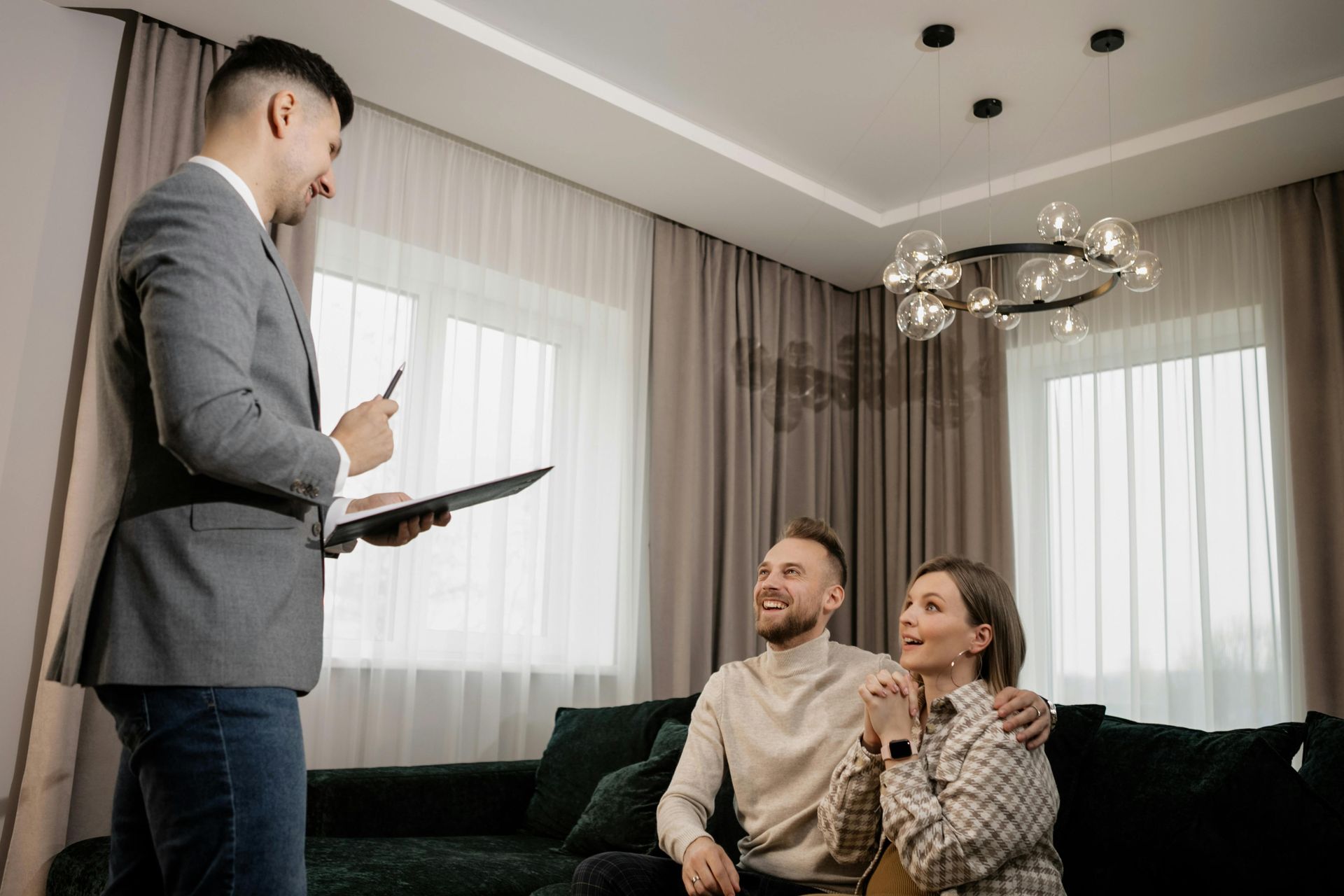 A professional in a gray blazer stands holding a clipboard, speaking to a smiling couple sitting together on a couch.