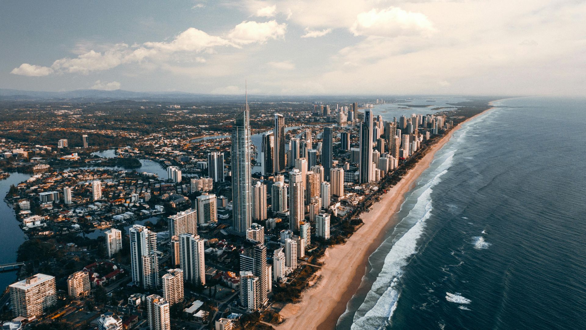 Aerial view of the Gold Coast skyline with tall skyscrapers bordering a long sandy beach and the Pacific Ocean.