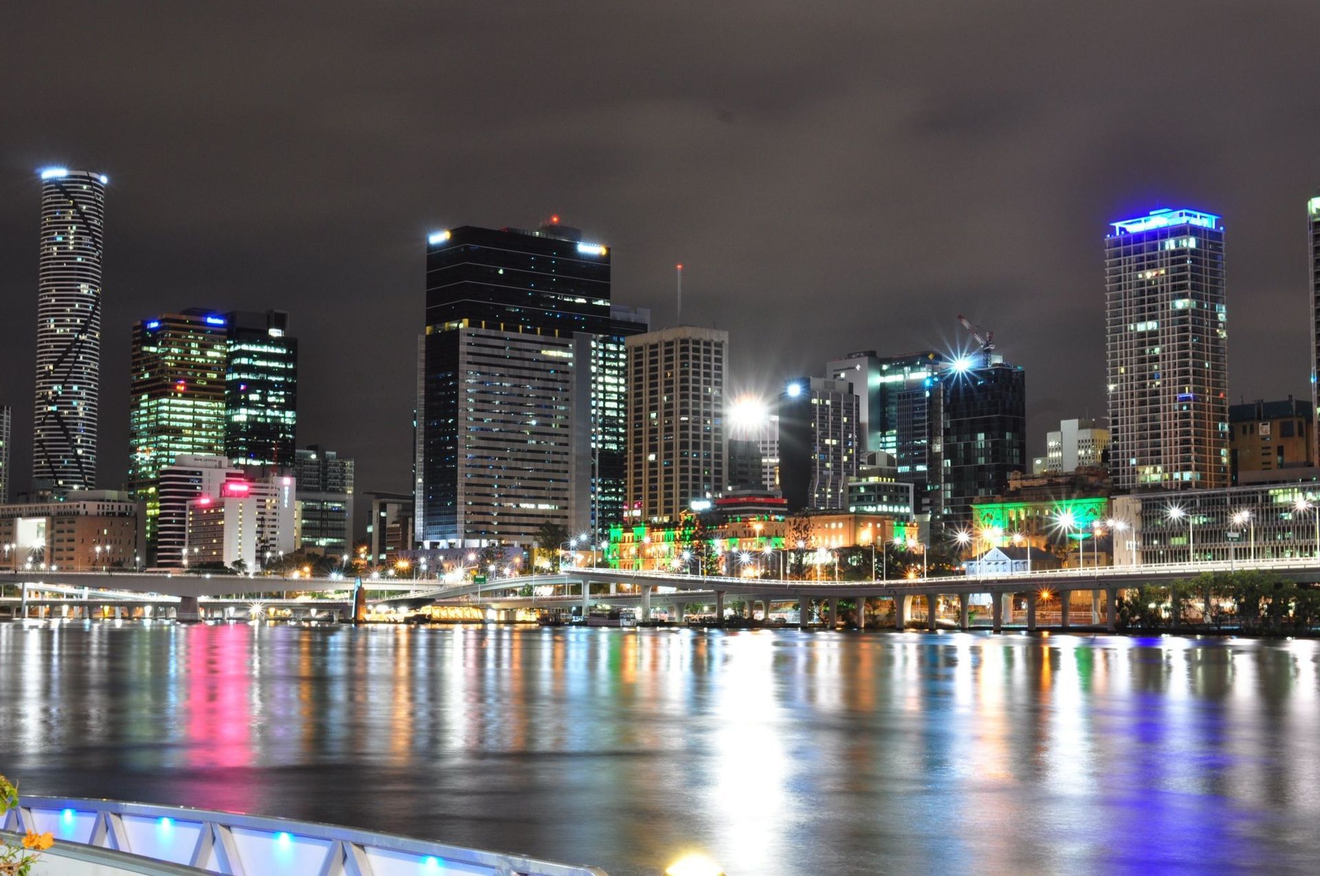 City skyline at night reflecting over a river, with illuminated skyscrapers against a dark, cloudy sky.