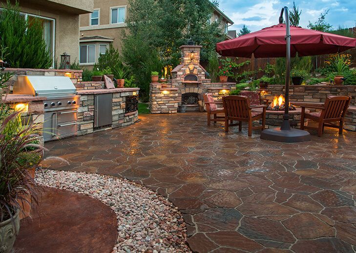 Outdoor patio with a stone grill island, fireplace, and seating area under a red umbrella at dusk.