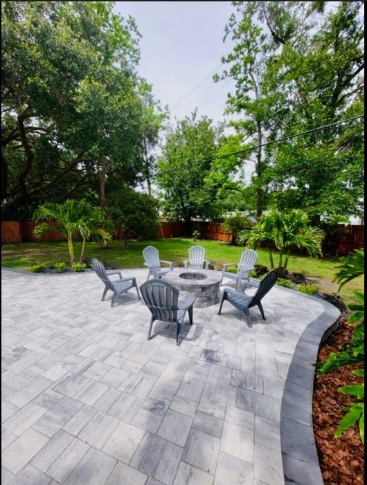 A circular stone fire pit sits on a light gray paver patio surrounded by six dark Adirondack chairs in a backyard.