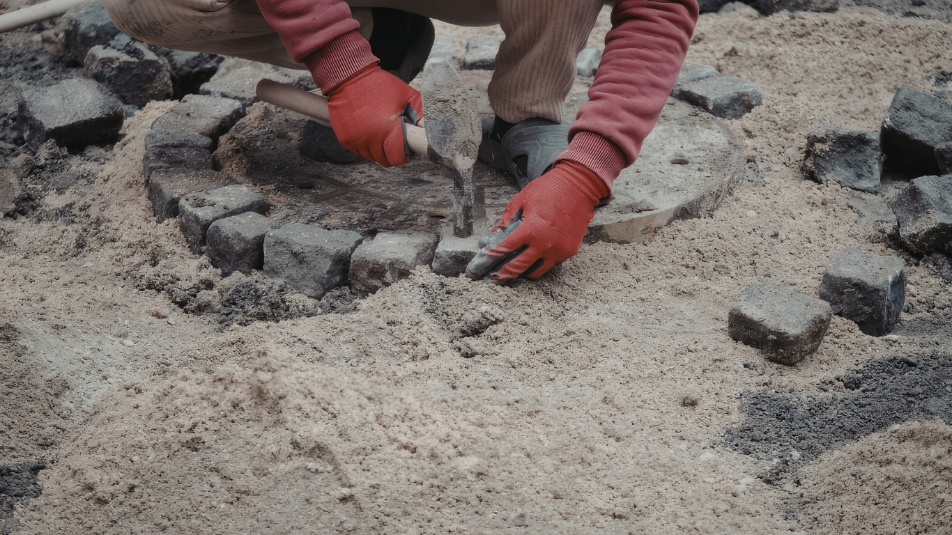 A person wearing red gloves uses a small hammer to set stone pavers into a circular pattern in a sand bed.