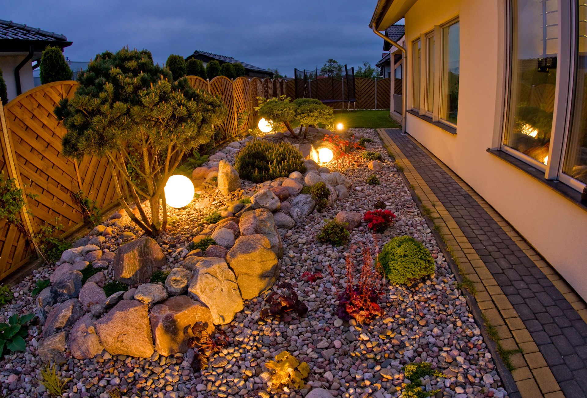 A landscaped garden bed at twilight with rocks, small shrubs, and glowing orb-shaped lights along a paved walkway.