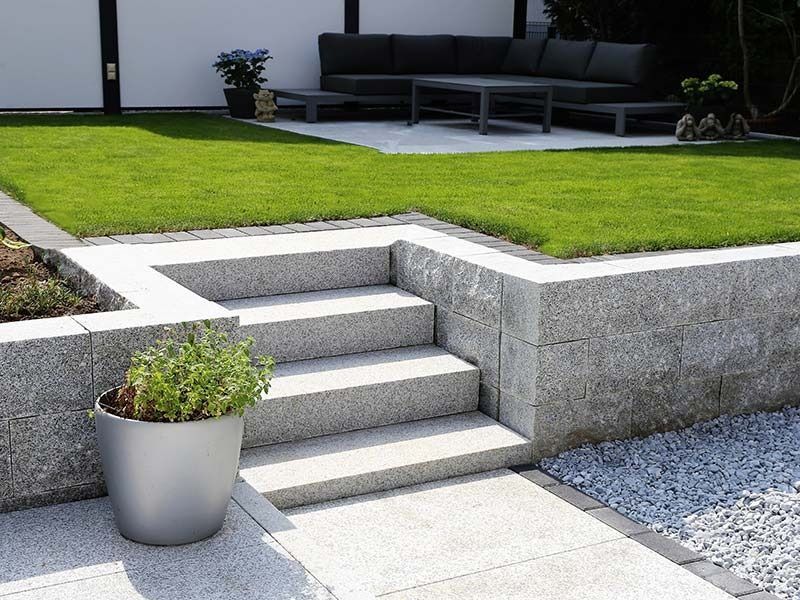 Granite steps leading up from a paved patio to a lawn with a sofa set, bordered by a stone wall and a potted plant.