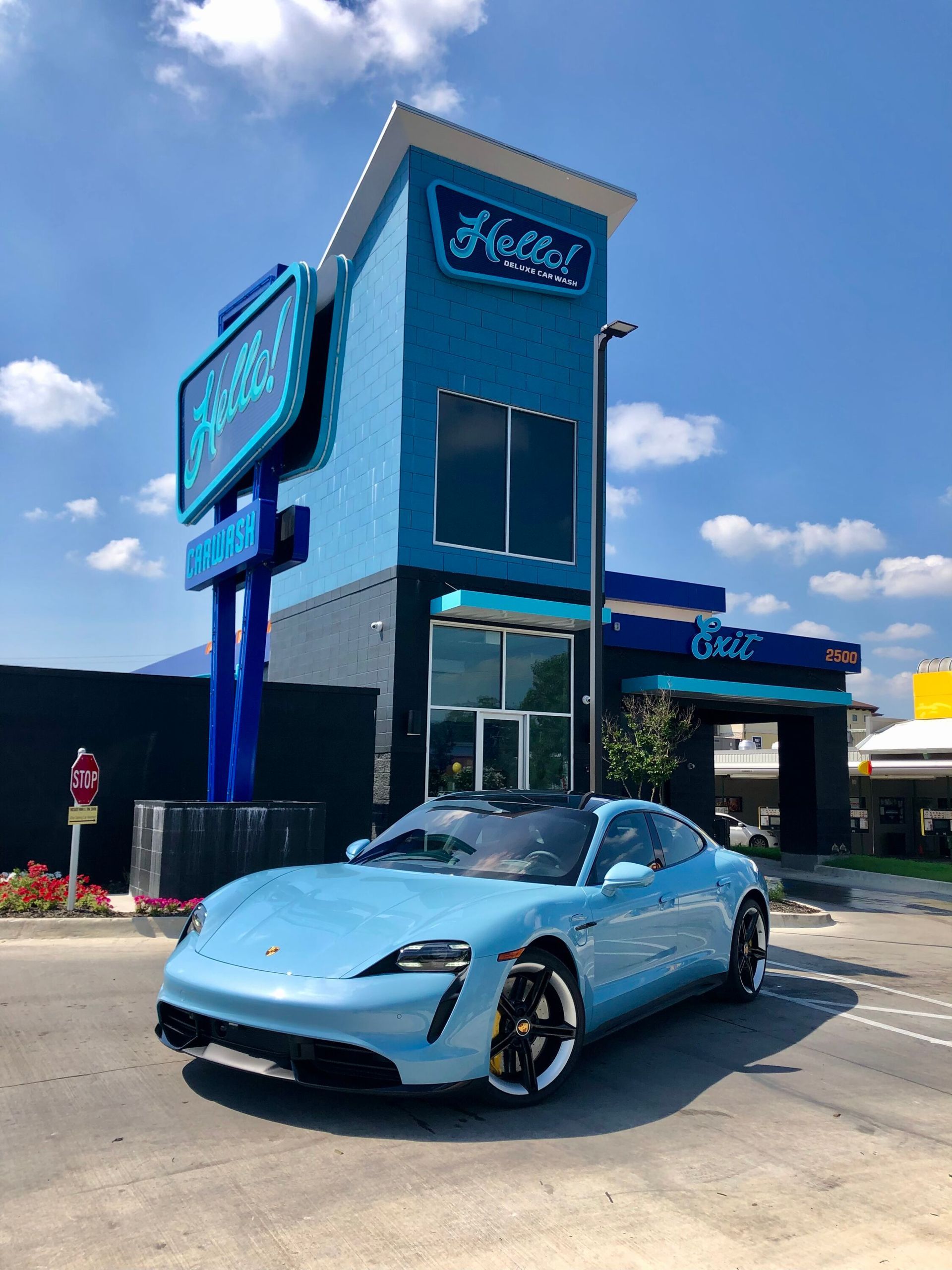 Light blue Porsche parked in front of a teal car wash building on a sunny day.