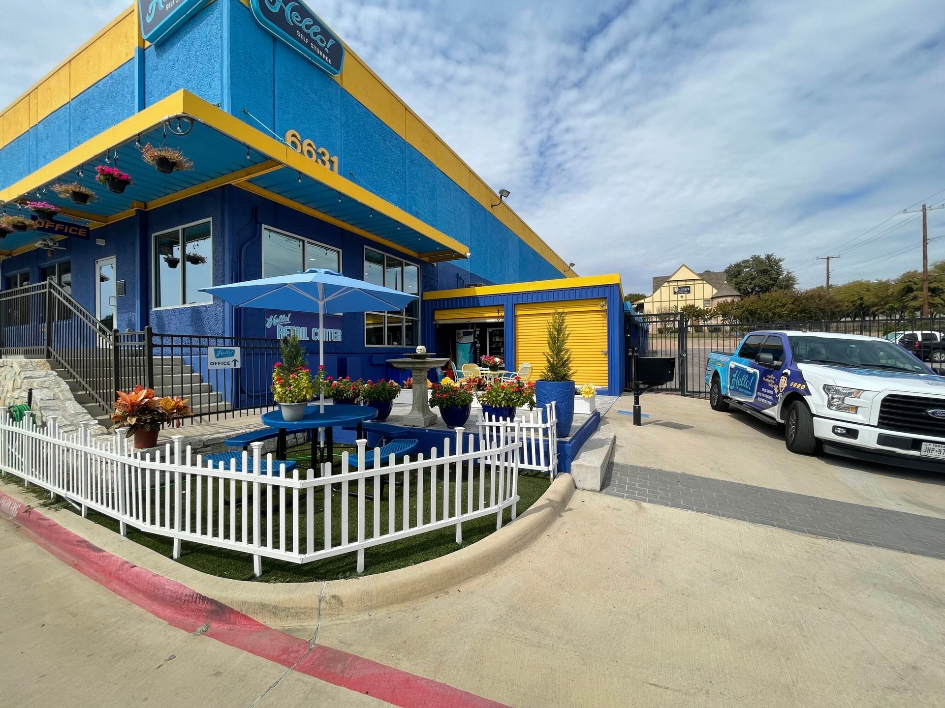 Blue and yellow building with a white picket fence and a blue umbrella-covered table. A truck is parked to the right.