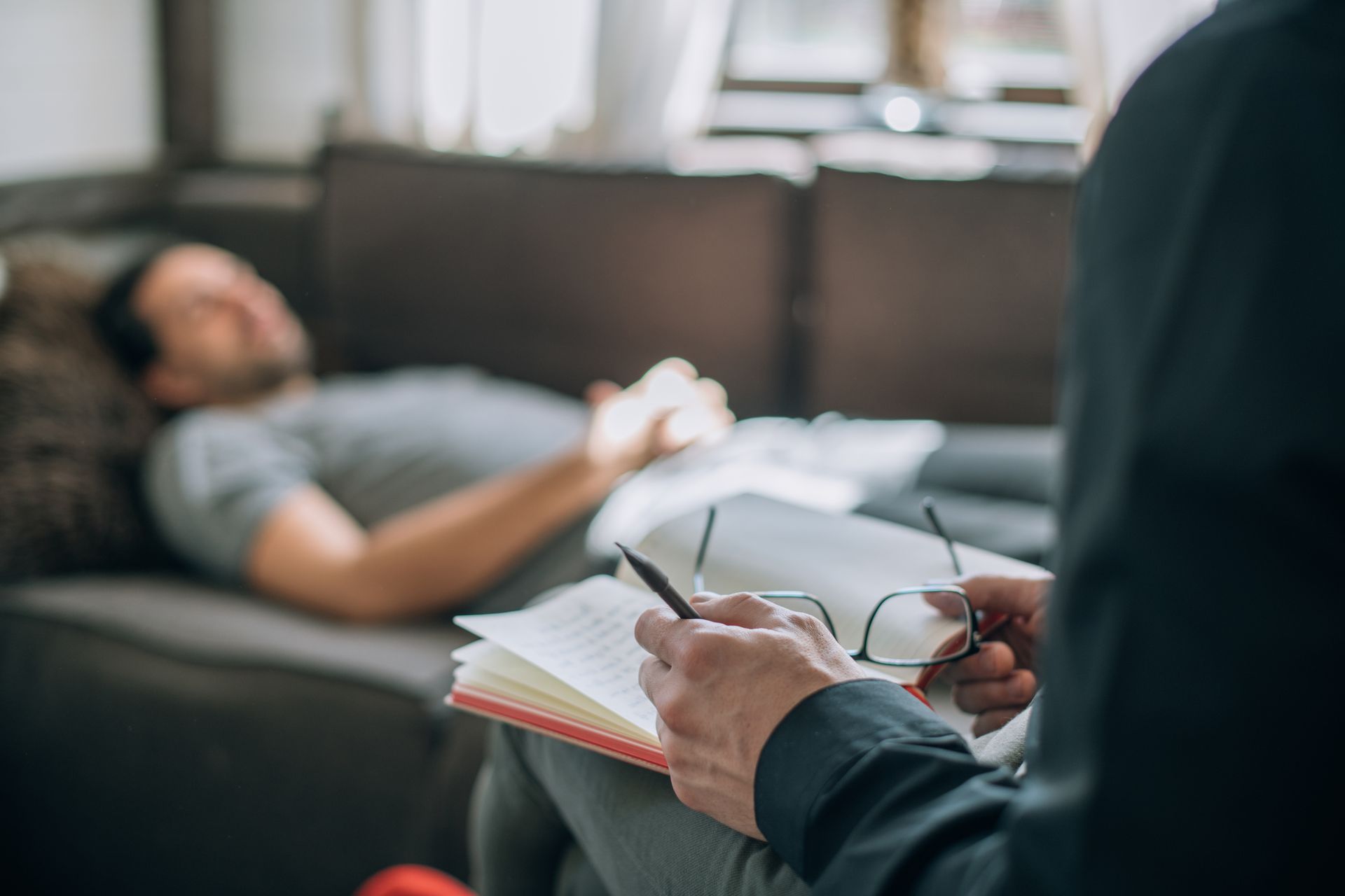 A man is laying on a couch while a man takes notes in a notebook.