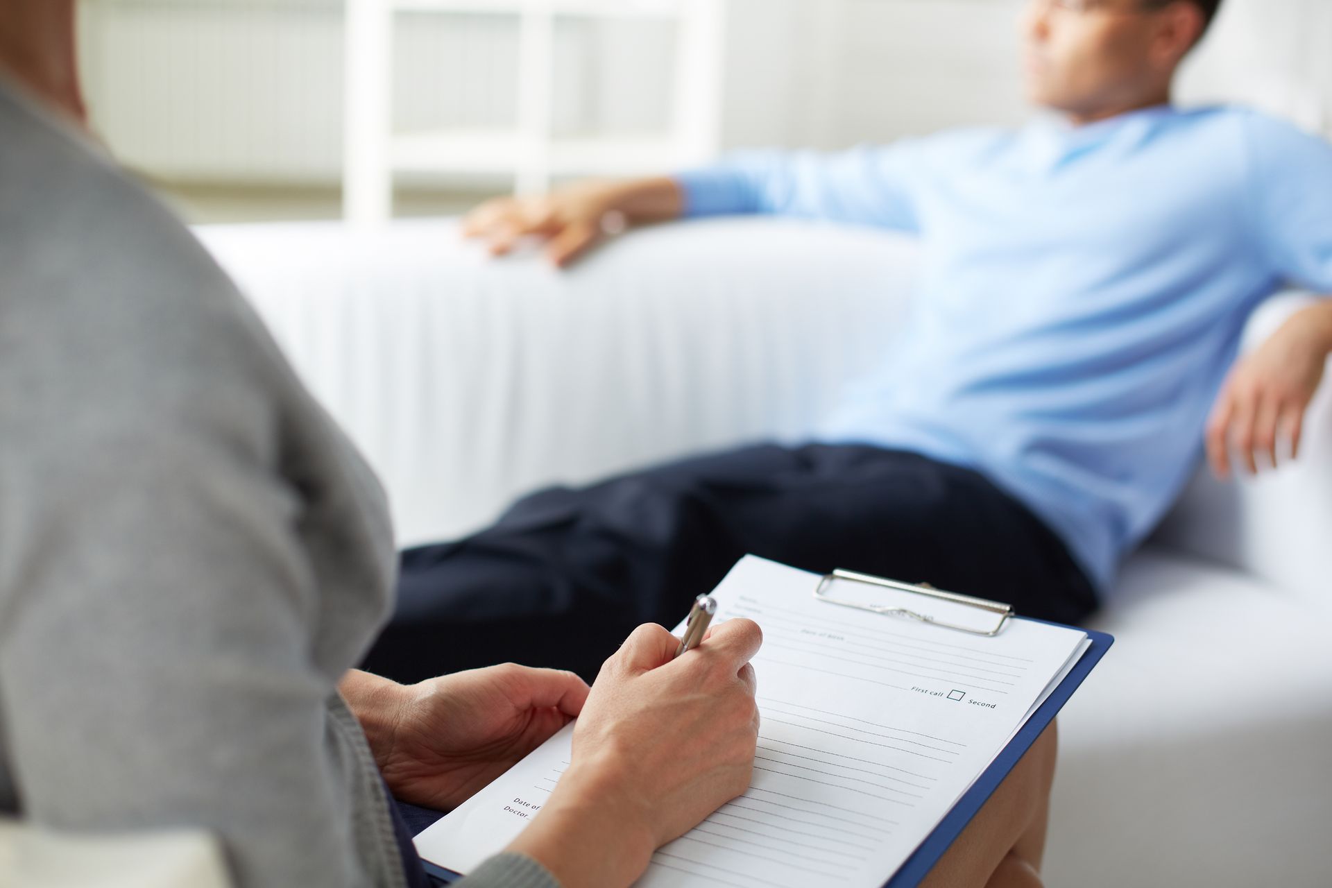 A man is laying on a couch while a woman writes on a clipboard.