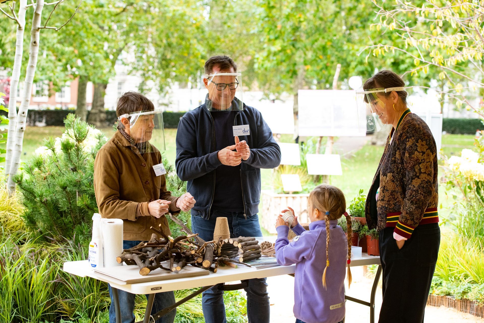 People at a table with crafts, wearing face shields, outdoors. Child building with wood pieces.