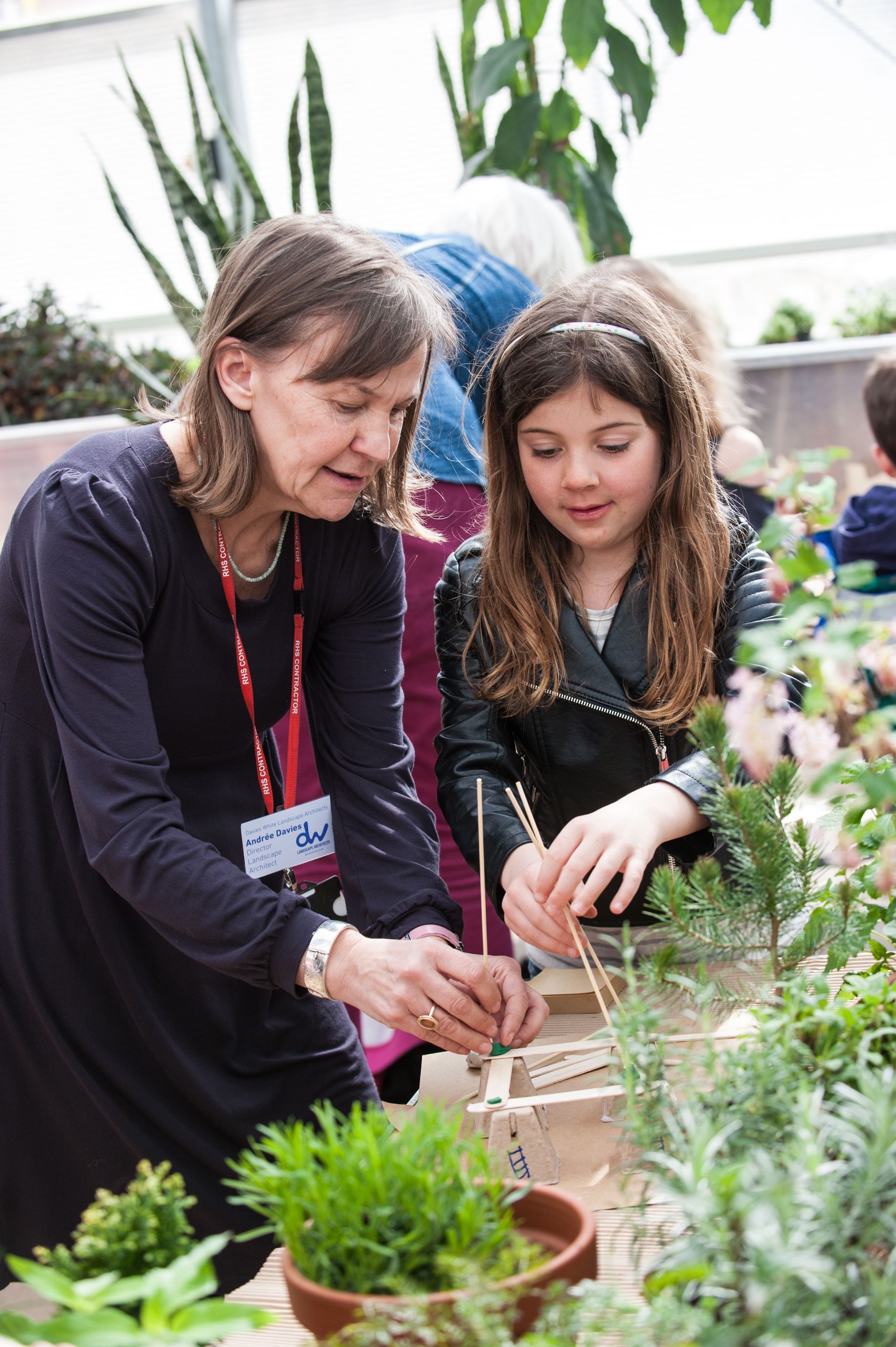 Woman and young girl building with sticks and plants in a greenhouse, smiling.