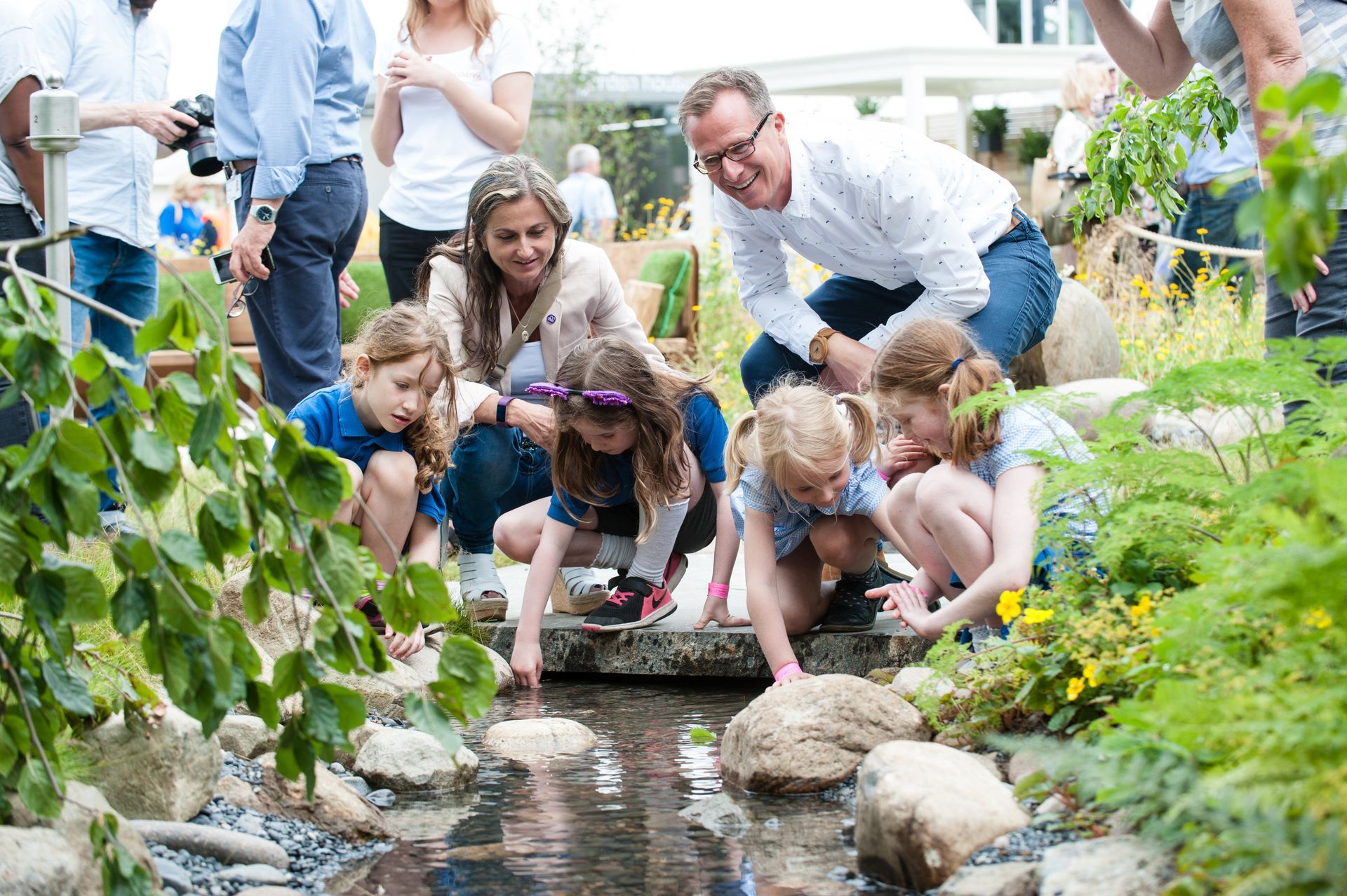 Children and adults explore a stream, reaching into the water amidst greenery.
