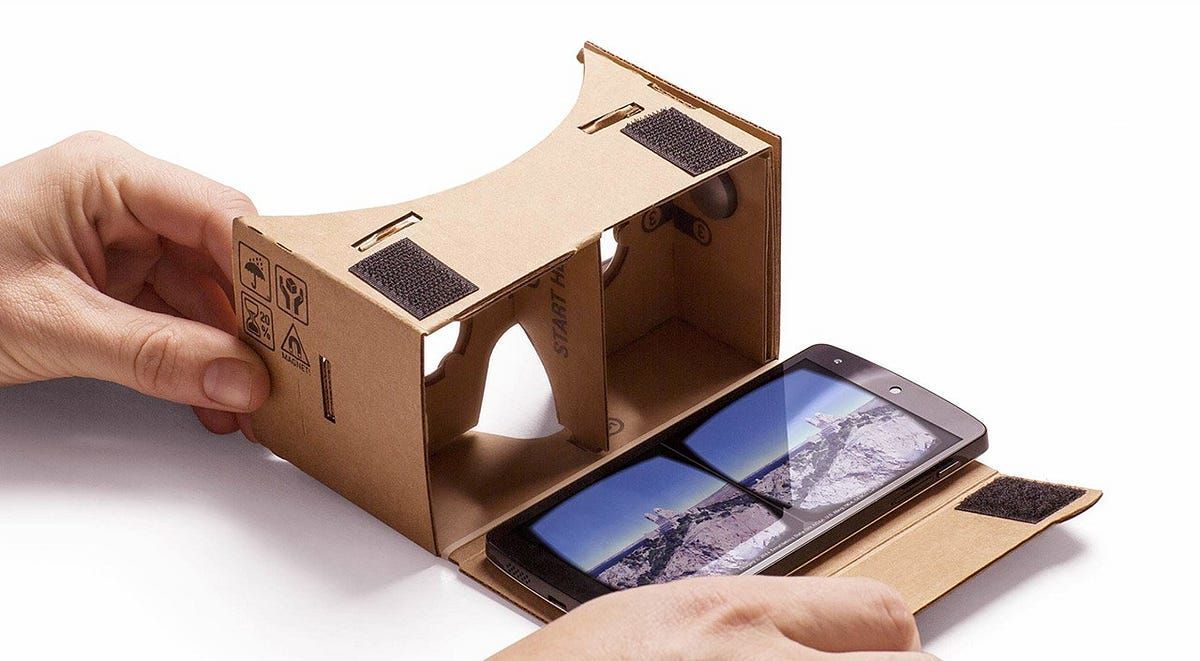 Person holding a cardboard VR viewer with a smartphone showing a split-screen mountain view.