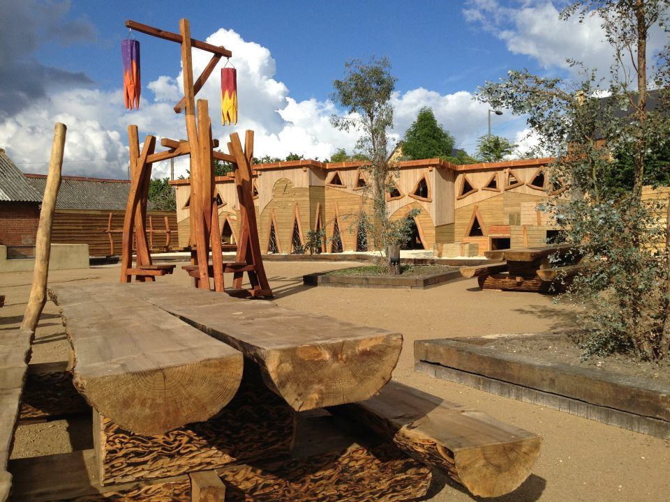 Wooden playground with bench, structure, and colorful flags; backdrop is tan building with openings and blue sky.