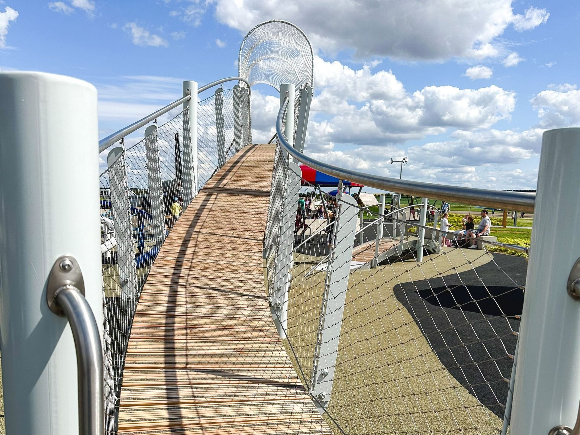 Playground bridge with wooden planks, metal supports, and netting, against a blue sky with clouds.