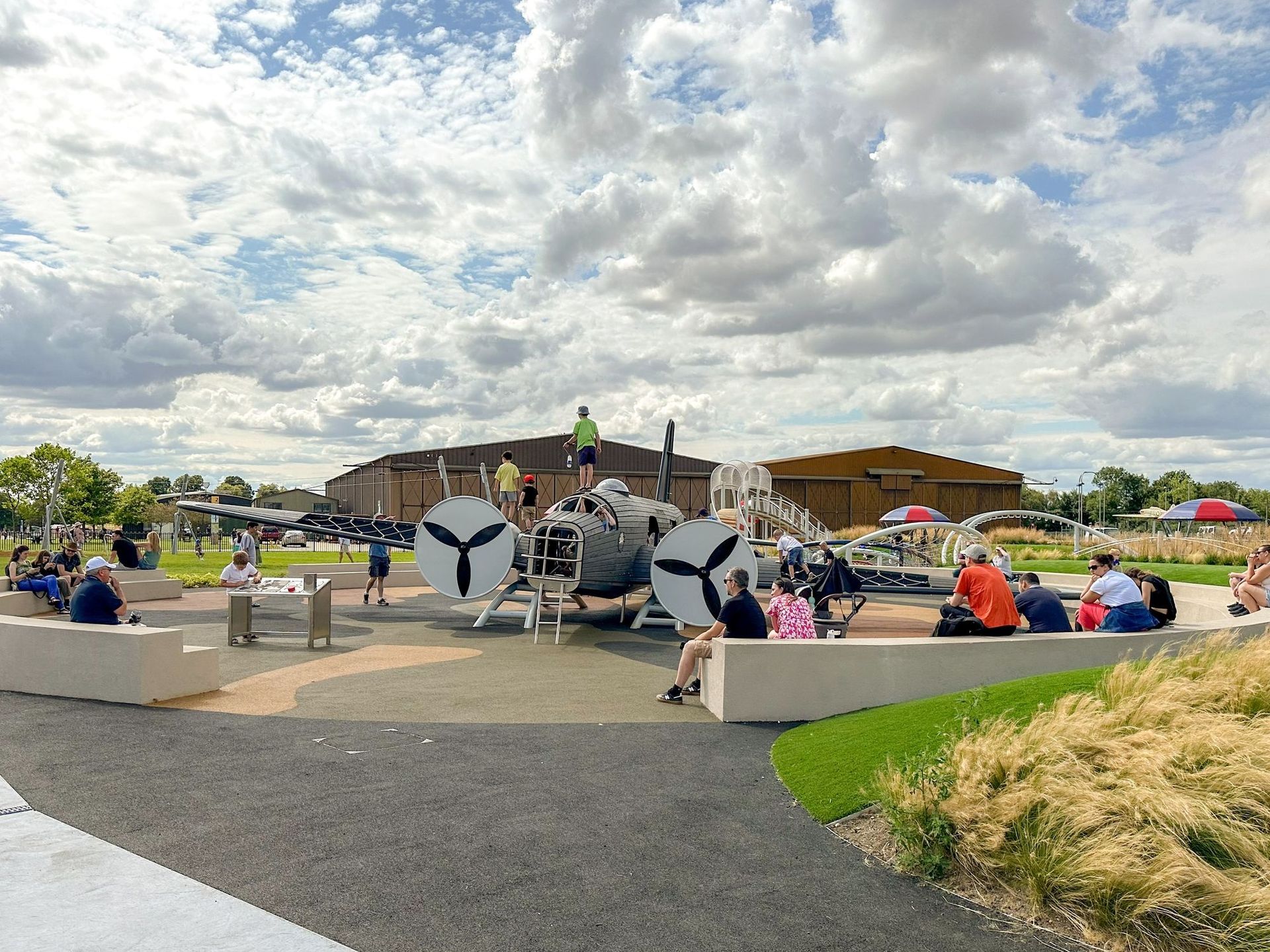 Playground with large airplane-themed structure; people sitting, playing; cloudy sky.