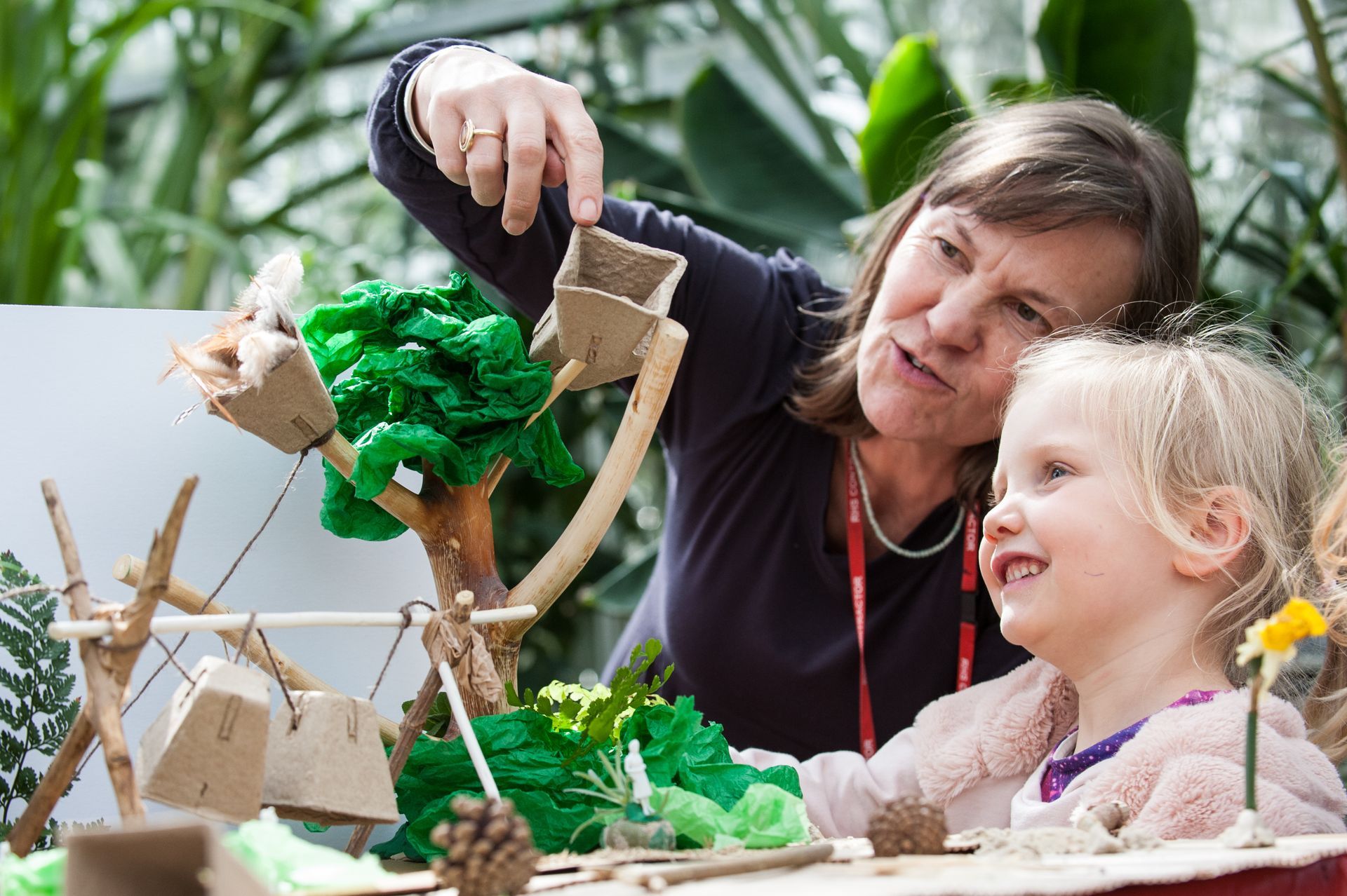 Woman points at a craft water feature, showing it to a smiling young child. Indoors, green plants in background.